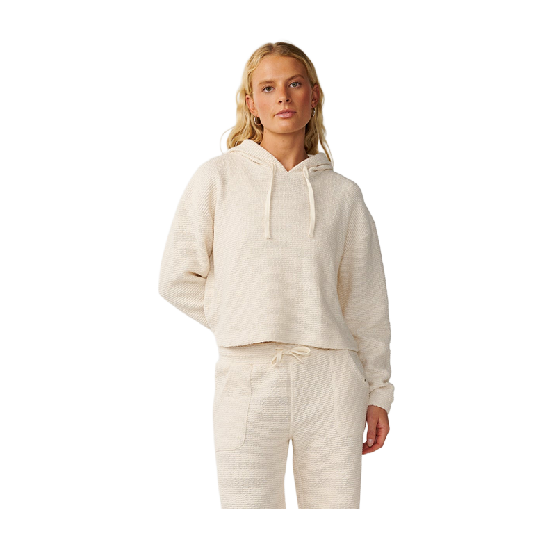 A woman modeling a matching cream-colored textured loungewear set, featuring a cropped pullover hoodie with drawstrings and coordinating jogger pants, shot against a solid white background.