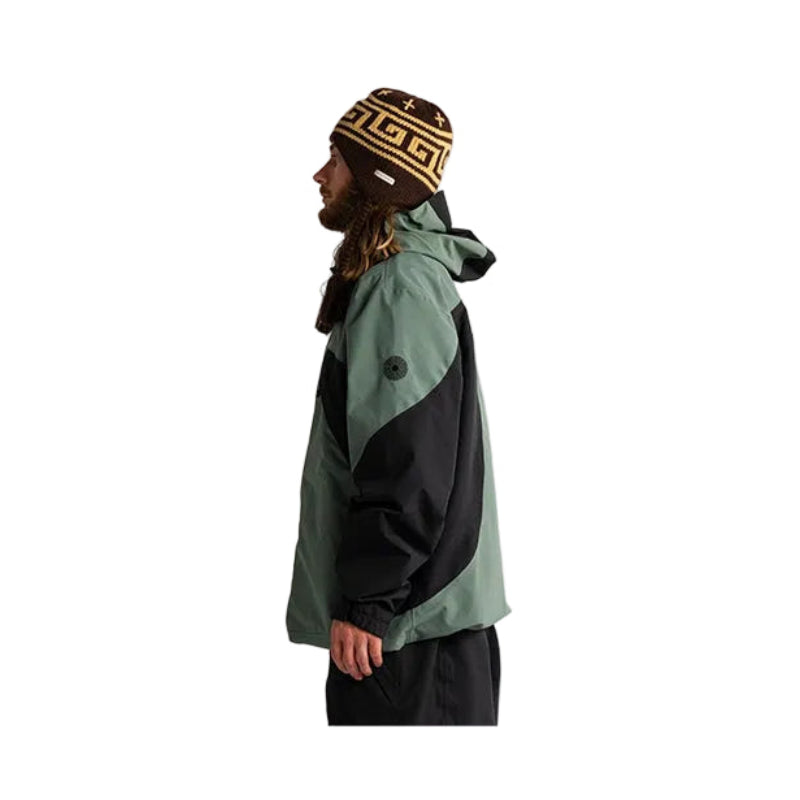 A man in profile wearing a green and black color-blocked hooded jacket, a brown and gold Greek key pattern beanie, and black pants, standing against a plain white background.