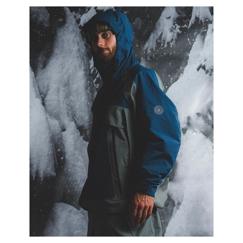 A man wearing a blue and gray hooded technical jacket stands in front of a snowy, icy outdoor background. The jacket has a blue upper section, gray lower panels, and a circular logo patch on the left sleeve.