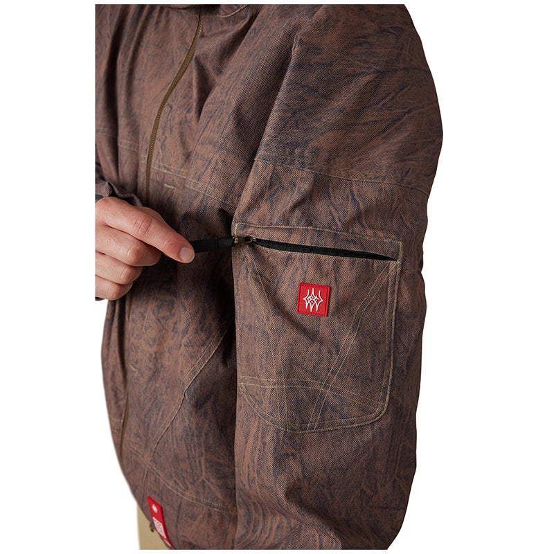 Close-up of a brown textured jacket with a hand pulling a black zipper on the sleeve pocket, featuring a small red brand patch.