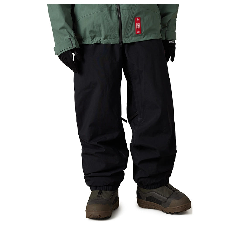 A person wearing black snow pants, a muted green jacket, black gloves, and brown snowboard boots, shown from the waist down on a plain white background.