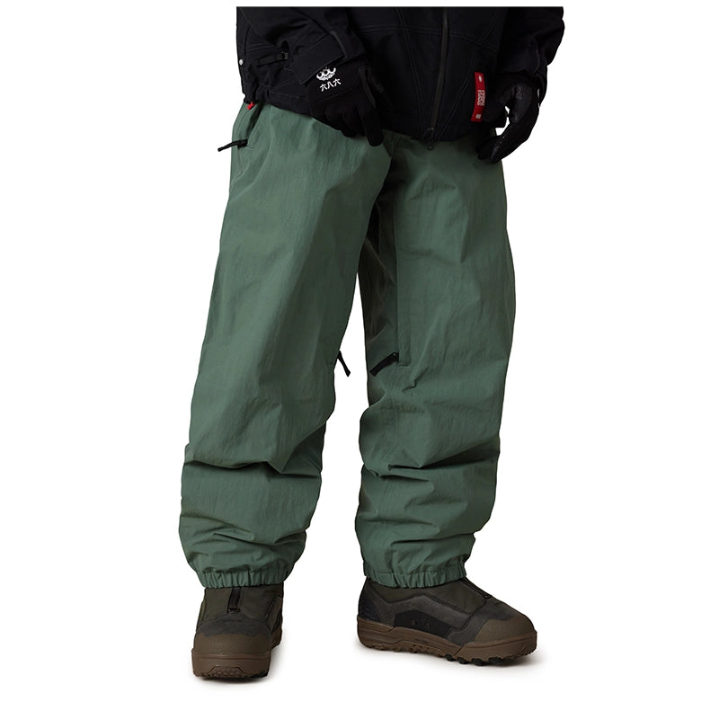 A person wearing dark green snow pants with elastic cuffs, paired with brown snowboard boots and a black jacket, shown from the waist down on a white background.