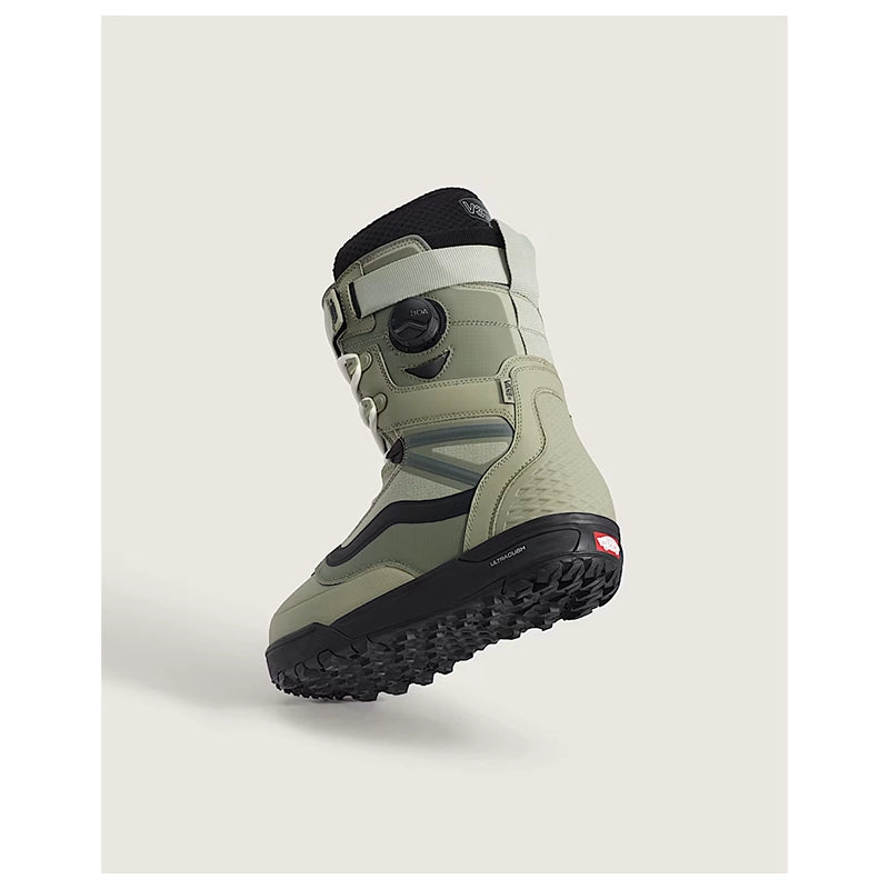 A single snowboard boot floating against a light warm gray background. The boot is primarily sage green with black and white details, featuring a dial closure system and a textured black sole.