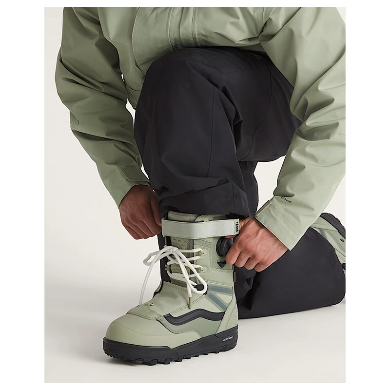 A close-up of a person wearing an olive green ski jacket and black pants, adjusting the cuff of a light olive green ski boot with white laces.