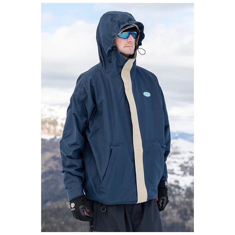 A man wearing a navy blue hooded ski jacket with a beige center stripe and a small oval logo patch on the chest, standing outdoors in a snowy mountain setting.