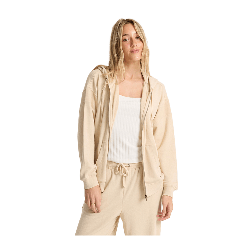A woman modeling a light beige zip-up hoodie and matching sweatpants over a white ribbed tank top, photographed against a solid white background.