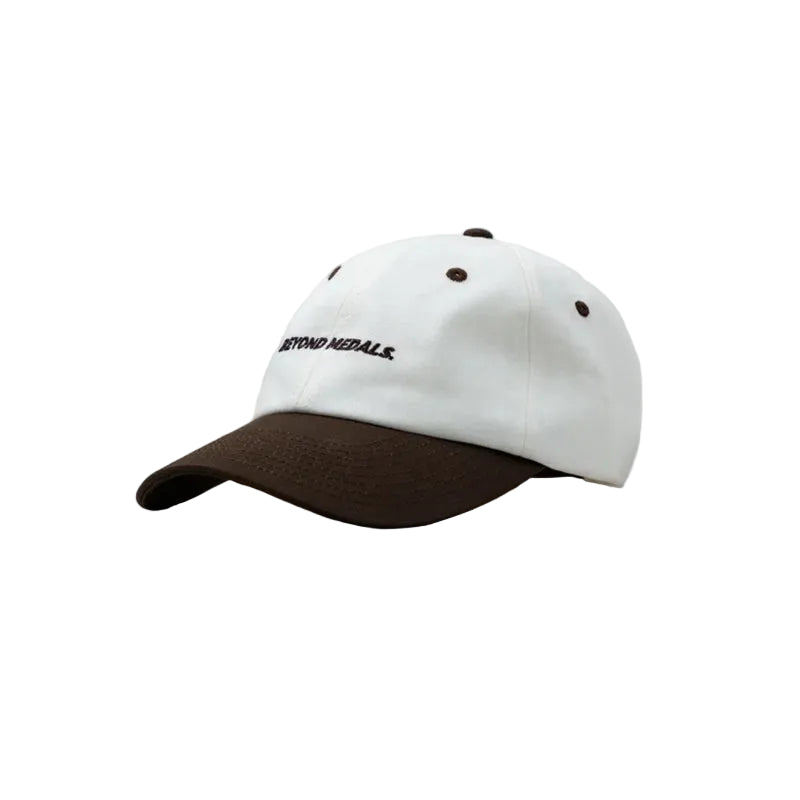 A baseball cap with a white crown and a dark brown visor, featuring the embroidered text 'BEYOND MEDALLS' in black on the front panel. The cap is photographed from a three-quarters angle against a solid light gray background.
