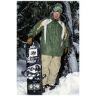 A man standing in the snow wearing a green and white ski jacket, beige pants, and a green beanie with goggles, holding a black snowboard with visible BCa Sports and Megathick branding.