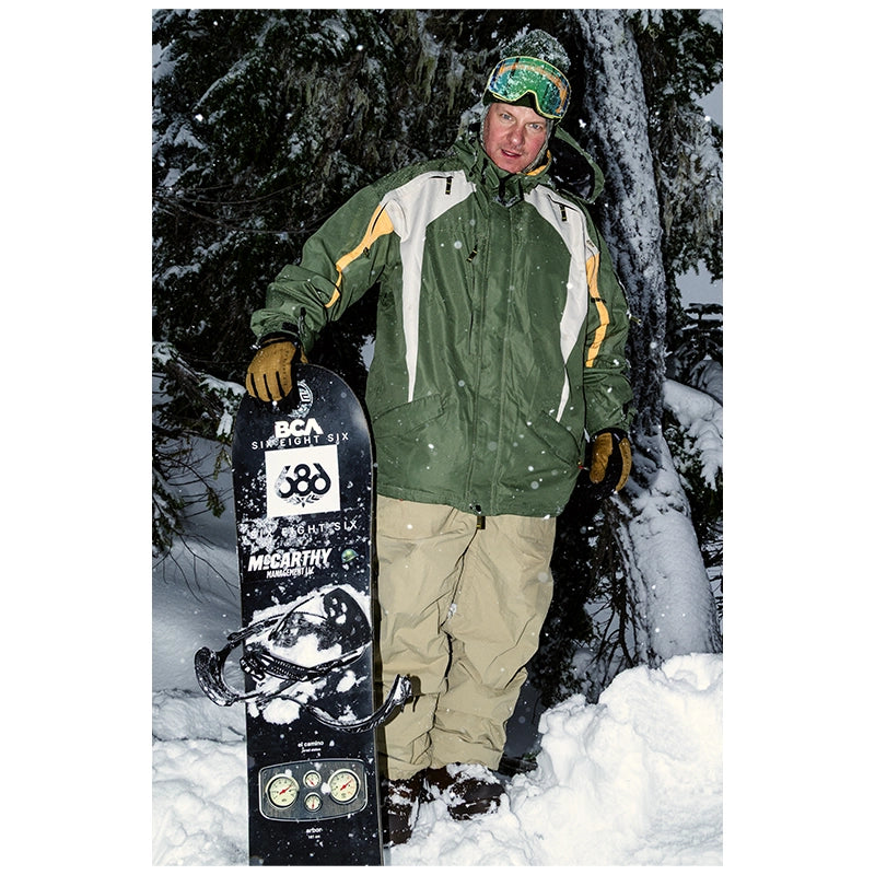 A man standing in the snow wearing a green and white ski jacket, beige pants, and a green beanie with goggles, holding a black snowboard with visible BCa Sports and Megathick branding.