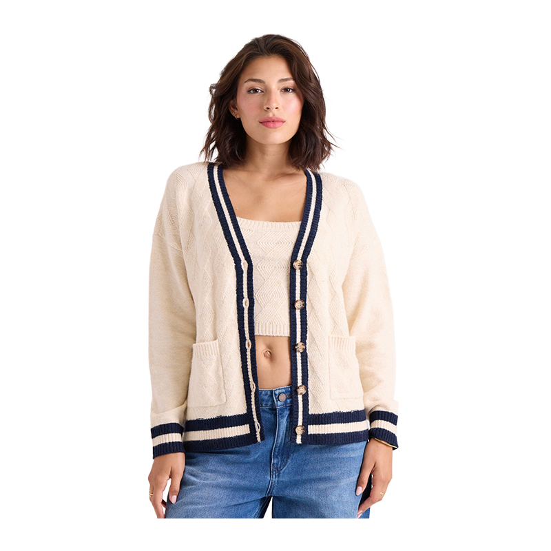 A woman modeling a cream cable-knit cardigan with navy blue trim and wooden buttons, paired with a matching cream crop top and blue jeans, against a solid white background.