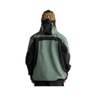 A person viewed from behind wearing a hooded jacket with a sage green body and black sleeves, hood, and shoulder panels, along with a brown and black patterned beanie, against a plain white background.