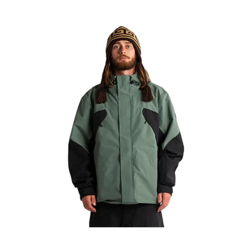 A man wearing a sage green and black technical jacket with a striped beanie, standing against a plain white studio background.