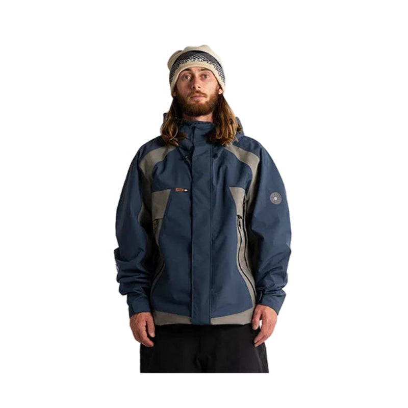 A man wearing a navy blue and gray technical outdoor jacket, a gray knit beanie, and black pants, standing against a plain white studio background.