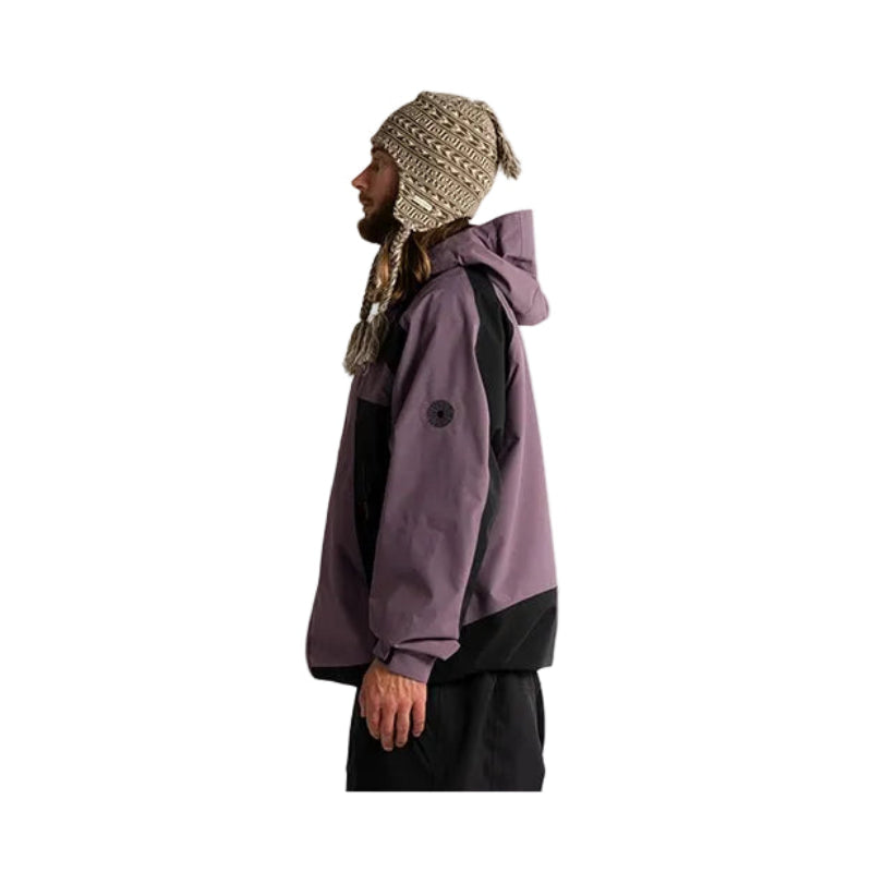 A man in profile wearing a two-tone purple and black hooded jacket, a beige knit beanie with ear flaps, and a patterned neck gaiter, standing against a plain white background.