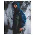 A man wearing a blue and gray hooded technical jacket stands in front of a snowy, icy outdoor background. The jacket has a blue upper section, gray lower panels, and a circular logo patch on the left sleeve.