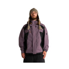 A man wearing a purple and black ski jacket, a patterned knit beanie, and black pants, standing against a plain white studio background.