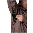 Close-up of a man wearing a brown camouflage-patterned jacket, opening a side zipper to reveal a black inner lining, against a plain white background.