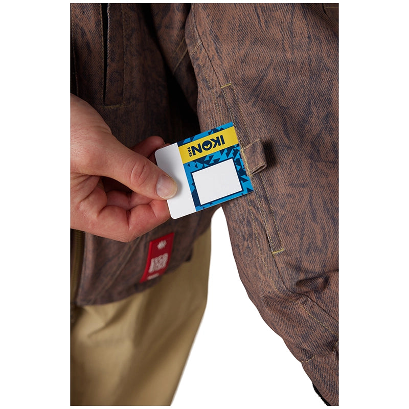 A close-up of a person's hand holding a small blue and yellow Koni card next to a brown patterned jacket pocket, with tan pants visible below.