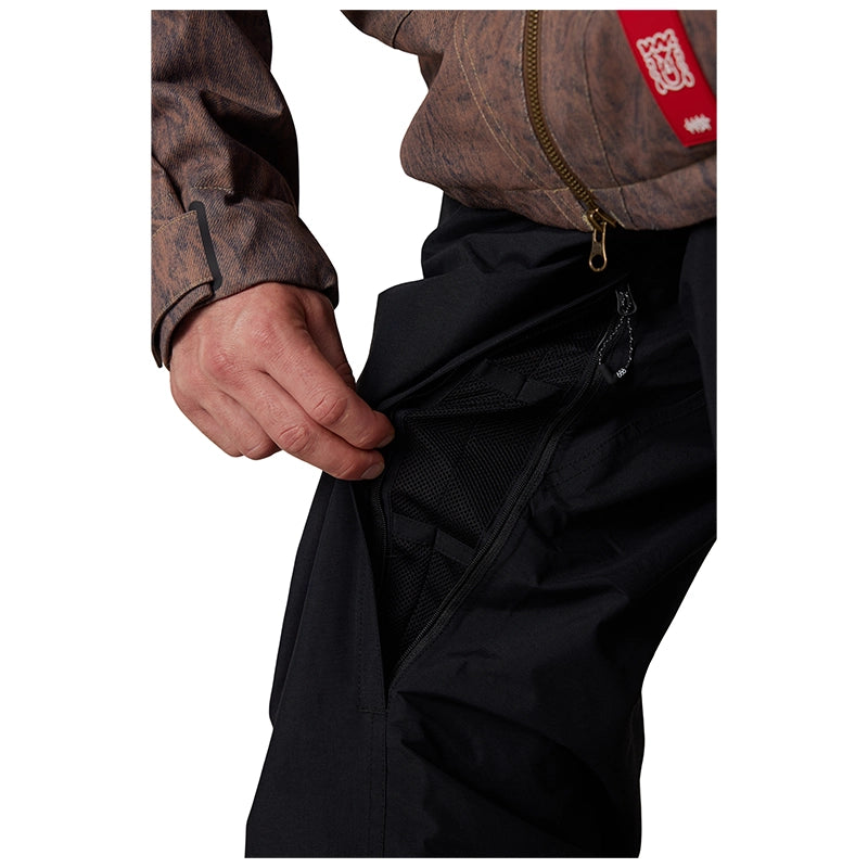 Close-up of a person wearing black pants and a brown patterned jacket, pulling open a side pocket of the pants to show the interior mesh lining. A red brand patch is visible on the jacket.
