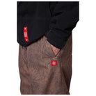 A close-up of a person wearing a black jacket and brown patterned pants, with a red rectangular logo patch visible on the jacket pocket and a matching patch on the pants.