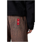 Close-up of a person wearing brown textured pants with a red rectangular patch on the back pocket and a black jacket, shown against a plain white background.
