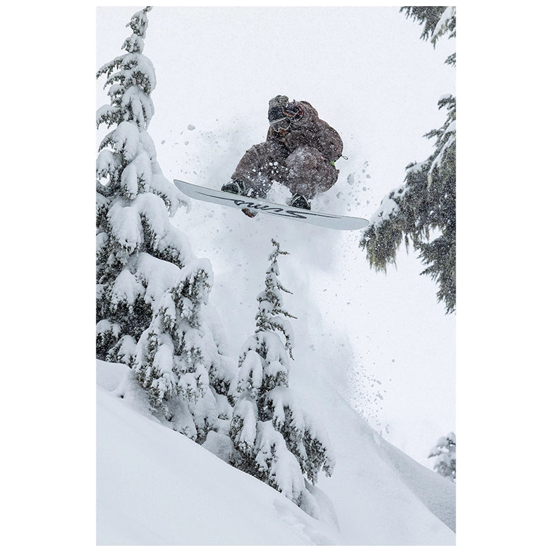 A snowboarder in brown and gray camouflage jumps through the air in deep snow, with snow-covered pine trees on both sides.