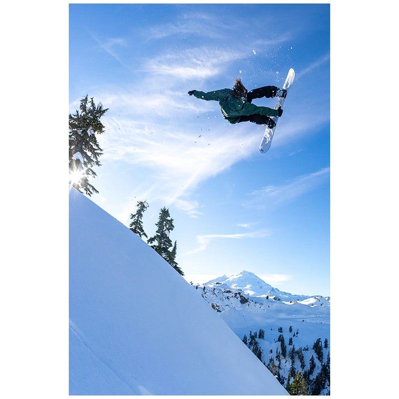 A snowboarder mid-air performing a jump in a snowy mountain setting under a bright blue sky with wispy clouds, with trees and a snow-covered slope visible.