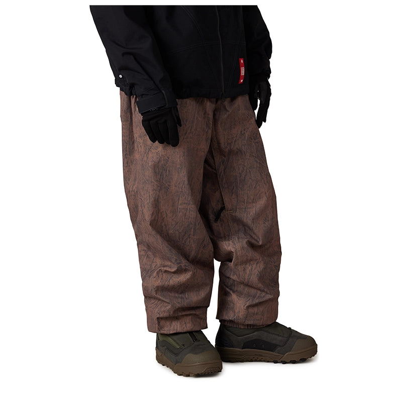 A person wearing brown patterned snow pants, a black jacket, and black gloves, shown from the waist down against a plain white background.