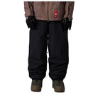 A person shown from the chest down wearing black snow pants, black gloves, a brown patterned jacket, and brown boots against a plain white background.