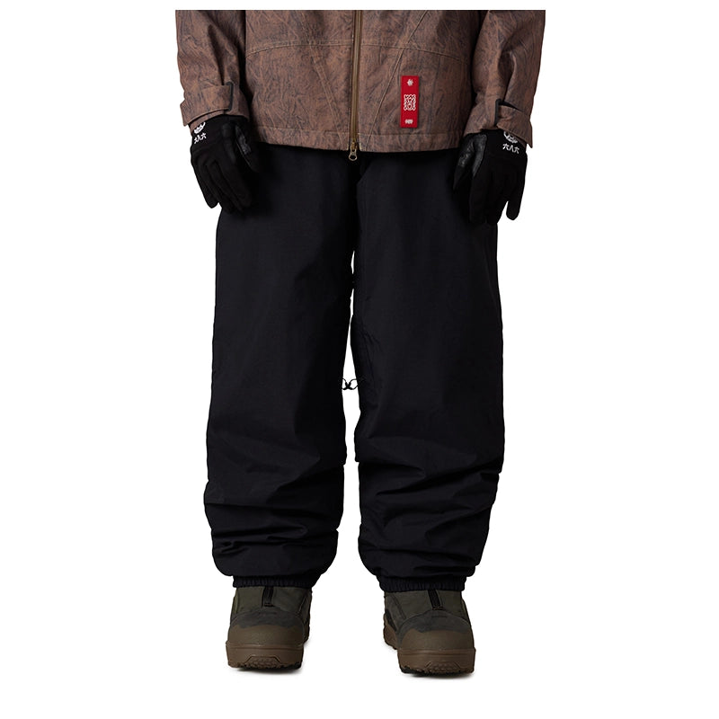 A person shown from the chest down wearing black snow pants, black gloves, a brown patterned jacket, and brown boots against a plain white background.