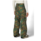 A person shown from the back wearing wide-leg camouflage cargo pants, a white long-sleeve top, and black-and-beige shoes against a light gray studio background.