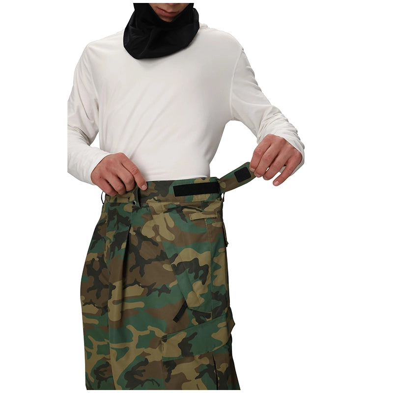 A model wearing a white long-sleeve shirt and a black neck gaiter, adjusting the waist of camouflage snow pants against a plain white background.