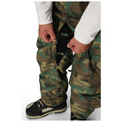 Close-up of a person wearing camouflage pants and black-and-white sneakers, pulling open a zipper on the pants with both hands.