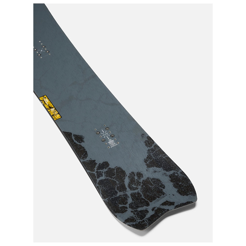 A close-up of a snowboard base showing the underside with a dark gray and black marbled pattern, a small yellow logo sticker, and a silver cross logo near the tail.