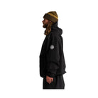 A man in profile wearing a black hooded jacket, black pants, and a brown knit beanie with gold trim, standing against a plain white background.