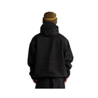 A person viewed from behind wearing a black hooded jacket, black pants, and a brown knit beanie with a yellow patterned band, against a plain white studio background.