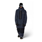 A model wearing a dark navy blue snowboard jacket with a black front panel, black snow pants, a black beanie, and goggles, standing against a plain white background.