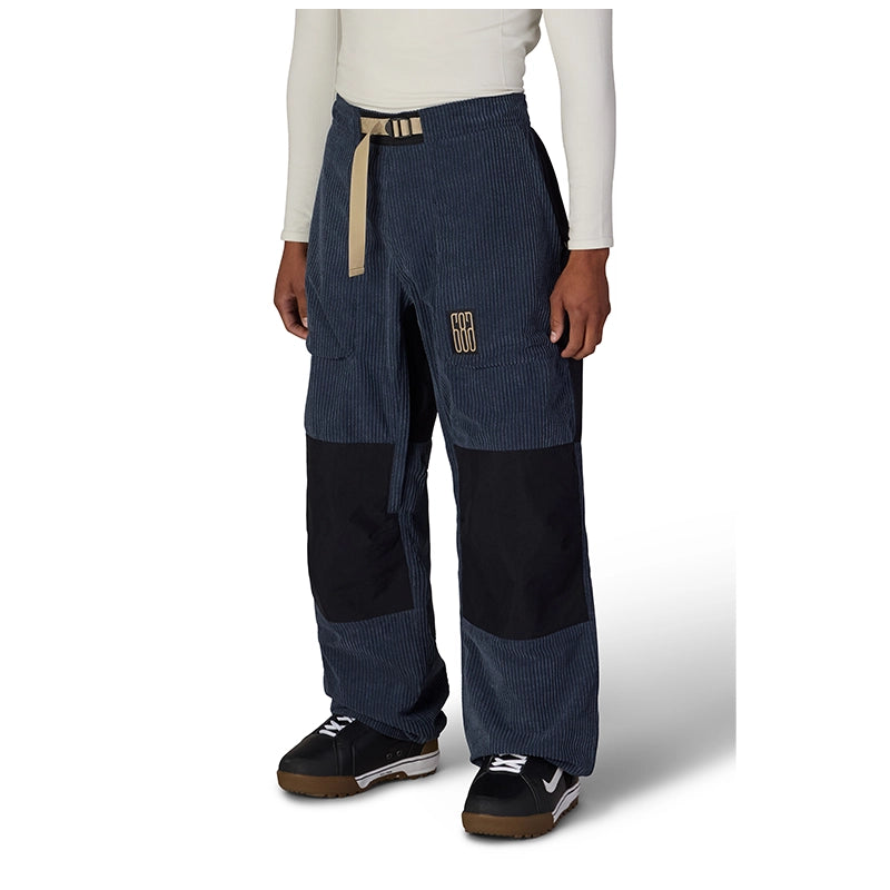 A person wearing wide-leg patchwork pants in dark blue corduroy and black, with a beige drawstring belt and a small gold logo patch on the thigh, paired with a white long-sleeve top and black sneakers.