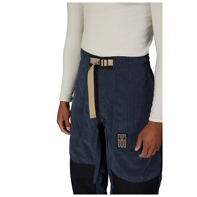 A person wearing dark navy corduroy pants with a beige belt and a gold geometric logo patch on the pocket, paired with a cream long-sleeve top against a white background.