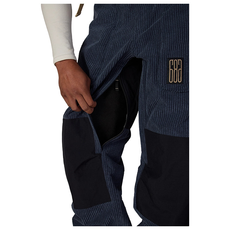 Close-up of a person wearing dark navy corduroy pants with black knee patches and a gold embroidered logo on the thigh, shown against a white background.