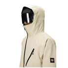 A side-profile view of a person wearing a light beige ski jacket with a high hood, black zipper details, and a black logo patch on the sleeve, set against a plain white background.