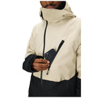 A close-up of a person wearing a hooded jacket in light beige and black, holding a black smartphone in a side zip pocket. The jacket has a high collar, a front zipper, and a contrasting black lower panel and sleeve cuffs, shown against a plain white background.
