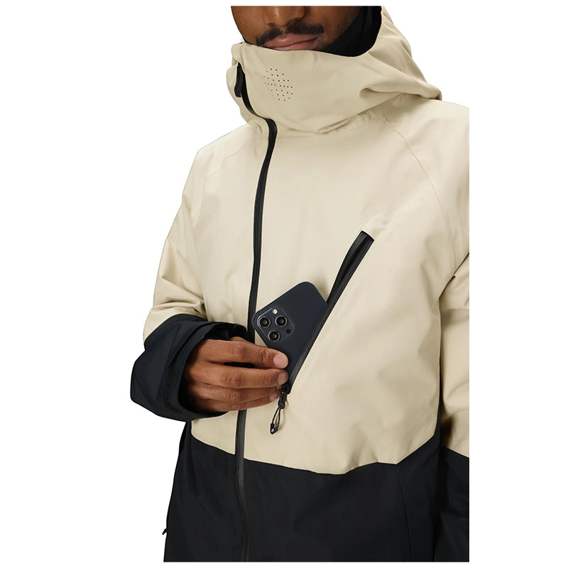 A close-up of a person wearing a hooded jacket in light beige and black, holding a black smartphone in a side zip pocket. The jacket has a high collar, a front zipper, and a contrasting black lower panel and sleeve cuffs, shown against a plain white background.