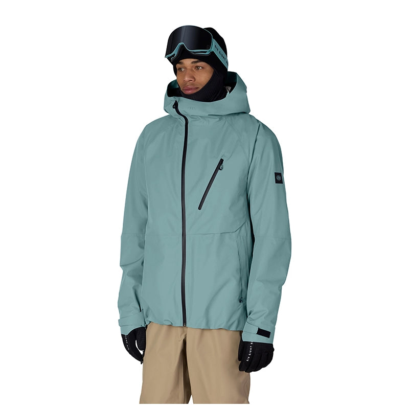 A man wearing a light teal ski jacket with black zippers, black gloves, a black neck gaiter, and ski goggles, standing against a solid white background.