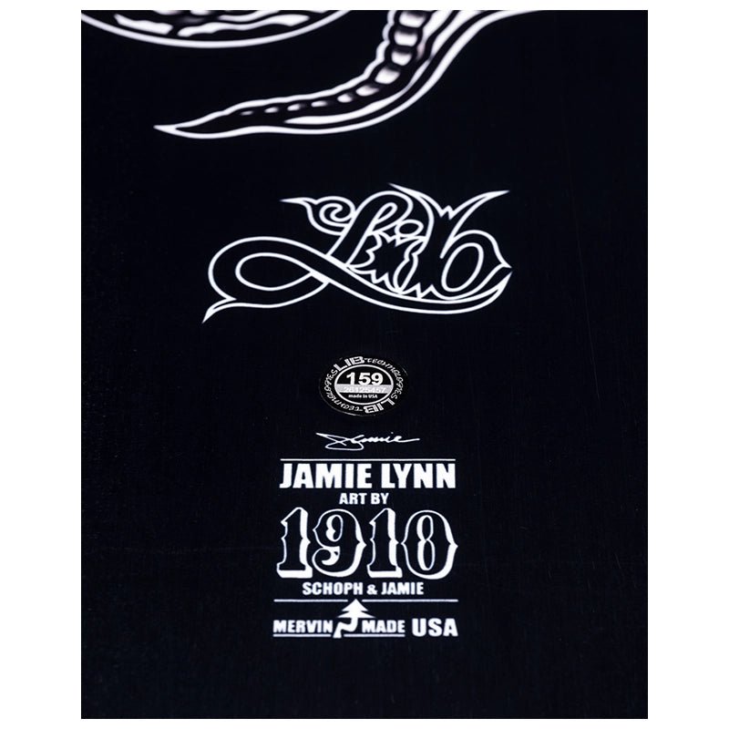A close-up of a black fabric surface featuring white printed graphics and text, including a stylized script logo, a circular emblem reading 159, and text that says Jamie Lynn Art by 1910, Schopf & Jamie, and Mervin Made USA.