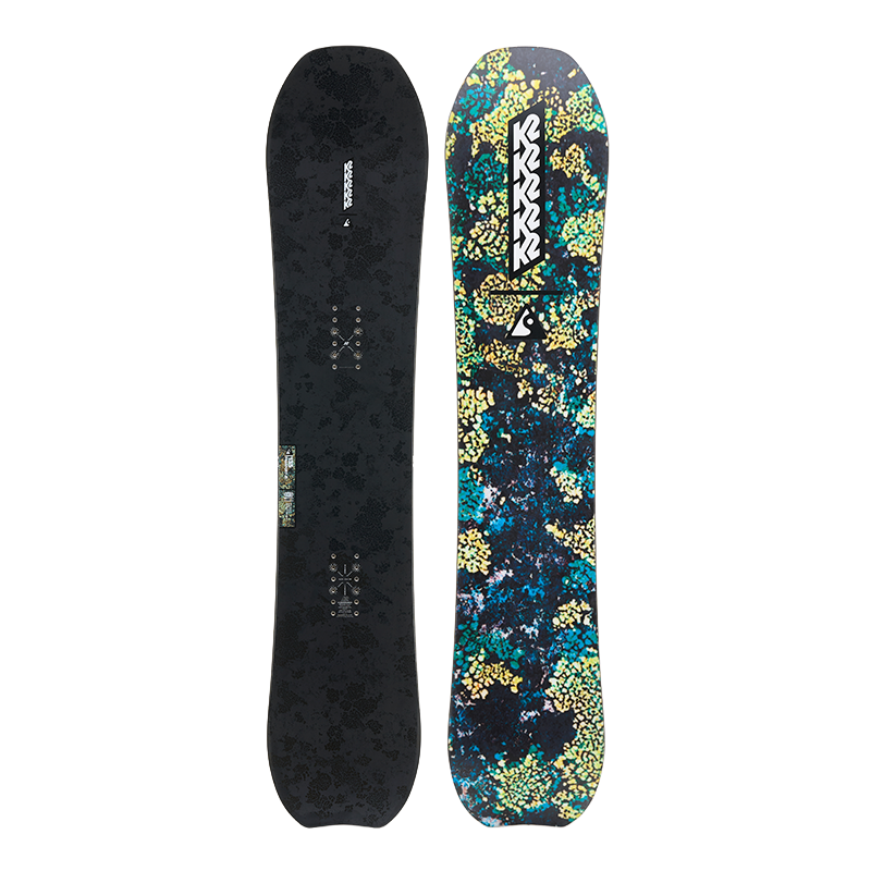 A snowboard shown from both sides against a plain white background. The left side shows a dark charcoal/black textured base with binding mount areas, and the right side shows a vibrant, multicolored floral pattern in teal, yellow, and dark blue with a white and black K2 logo.