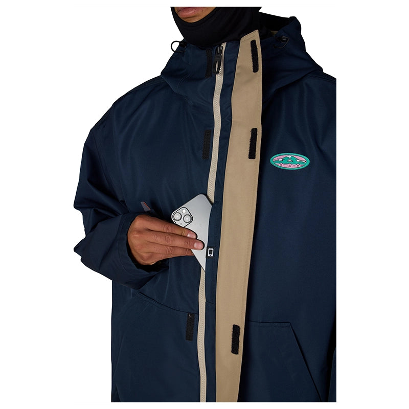 A close-up of a person wearing a navy blue hooded jacket with a beige front zipper panel and a small oval logo patch on the chest. The person is holding a smartphone in the jacket pocket, shown against a plain white background.