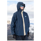 A man wearing a navy blue hooded ski jacket with a beige center stripe and a small oval logo patch on the chest, standing outdoors in a snowy mountain setting.