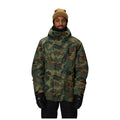 A man wearing a camouflage winter jacket, tan knit beanie, and black gloves, standing against a solid white background.