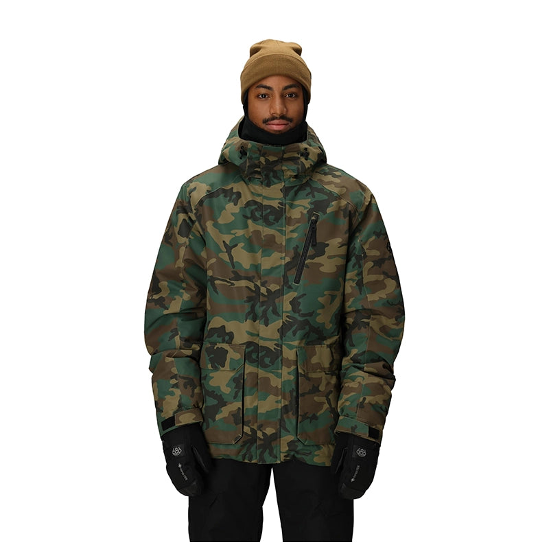A man wearing a camouflage winter jacket, tan knit beanie, and black gloves, standing against a solid white background.